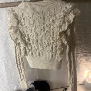 Vit stickad tröja med spetsdetaljer Zara  - Säljer en elegant vit stickad tröja med vackra spetsdetaljer och volanger. Tröjan har långa ärmar och en ribbad halsringning. Perfekt för en stilren look.