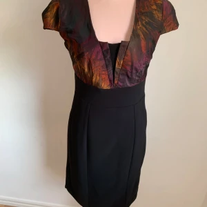 Flerfärgad klänning med svart kjol - Elegant klänning med en flerfärgad överdel i nyanser av röd, orange och lila. Kjolen är svart och figursydd för en smickrande passform. Klänningen har korta ärmar och en v-ringad halslinje som ger en stilren look.