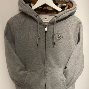 Burberry kofta - Snygg grå hoodie från Burberry med dragkedja och broderad logga på bröstet. Insidan av huvan har ett klassiskt rutigt mönster i beige, svart och röd. Perfekt för en stilren och bekväm look. M men passar S också. Helt ny , äkta, bra kvalité. Nypris 7499kr . Skriv för mer frågor! 