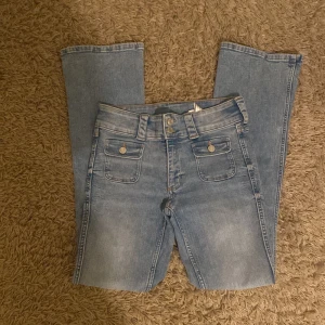 Blå bootcut jeans från H&M - Snygga blå bootcut jeans från H&M med justerbar låg midja. De har två framfickor med knappar och en klassisk jeanslook. Perfekta för en avslappnad stil.