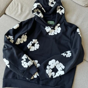 Denim tears hoodie - Säljer en svart hoodie med vita blommor. Den har en klassisk huva och långa ärmar. Perfekt för en avslappnad stil.