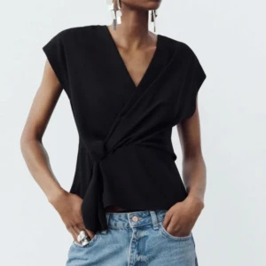 Svart topp med knytning - Snygg svart omlottopp med knytning i midjan. Toppen har en v-ringad design och är ärmlös, vilket ger en stilren och modern look. Perfekt för att matcha med jeans eller kjol. Kommer ej till användning längre!!💕