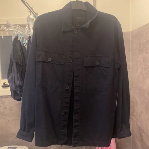 Svart overshirt med knappar - Säljer en stilren svart overshirt med knappar framtill. Jackan har långa ärmar och två bröstfickor med lock och knappar. Perfekt för lager-på-lager-stil och passar till många olika outfits. Aldrig använts därför säljer jag den.
