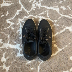 Svarta sneakers från Guess. - Snygga svarta sneakers från Guess med quiltad design och guldfärgade detaljer. Skorna har snörning och en vit sula som ger en stilren look. Perfekta för en casual stil. 