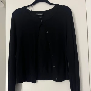 Svart kofta från Monki - Säljer en stilren svart kofta från Monki i storlek S. Koftan har långa ärmar och knappar framtill, perfekt för lager-på-lager-stil. Enkelt att matcha med olika outfits.