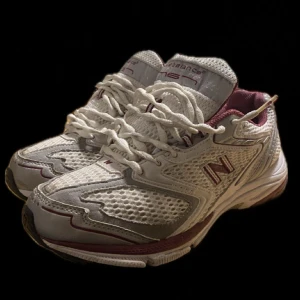 Vita och vinröda sneakers från New Balance - Snygga och bekväma vita sneakers från New Balance med vinröda detaljer. Skorna har en ventilerande meshdesign och klassisk snörning. Perfekta för en sportig look.