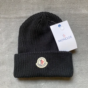 Svart stickad mössa från Moncler - Snygg svart stickad mössa från Moncler med broderad logotyp framtill. Perfekt för kyliga dagar och ger en stilren look. 