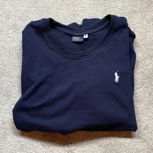 Mörkblå t-shirt från Ralph Lauren - Säljer en klassisk mörkblå t-shirt från Ralph Lauren med den ikoniska broderade loggan på bröstet. T-shirten är kortärmad och tillverkad i mjukt bomullsmaterial. Perfekt för en stilren och avslappnad look.