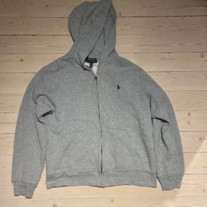 Grå hoodie från Ralph Lauren - Säljer en klassisk grå hoodie från Ralph Lauren med dragkedja och broderad logga på bröstet. Perfekt för en avslappnad stil. Den har långa ärmar och en bekväm huva.