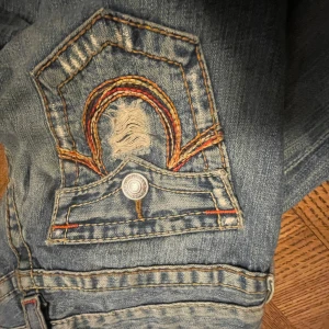 Blå jeans från True Religion - Säljer ett par blå jeans från True Religion med snygga broderade detaljer på bakfickorna. Jeansen har en bootcut-stil och lågmidjade 💗 säljer pågrund av att tyvärr är för små 😭35cm cm rakt över midjan och innerbenslängden är 85cm 💗
