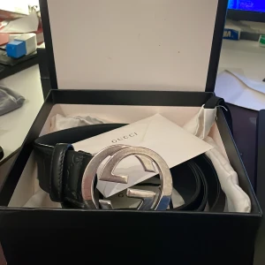 Svart präglat skärp från Gucci - Snyggt svart skärp från Gucci med en stor, rund spänne i silverfärg. Perfekt för att ge en lyxig touch till din outfit. Kommer i originalförpackning med Gucci-logga.