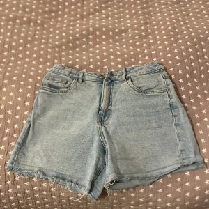 Ljusblå jeansshorts med fransar - Säljer ett par ljusblå jeansshorts med fransiga kanter. Shortsen har en klassisk femficksdesign och stängs med dragkedja och knapp. Perfekta för en avslappnad sommarlook!