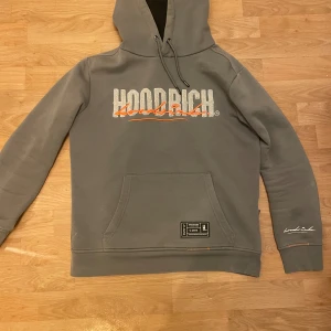 Grå hoodie från Hoodrich - Säljer en grå hoodie från Hoodrich med tryck i vitt och orange på både fram- och baksidan. Den har en stor ficka framtill och justerbar huva med dragsko. Perfekt för en avslappnad stil.