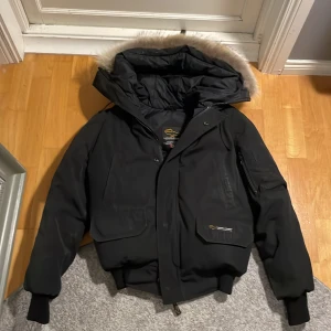 Svart dunjacka från Canada Goose - Säljer en svart dunjacka från Canada Goose med pälsfodrad huva och broderad logotyp på ärmen. Jackan har dragkedja och knappar. Skriv för fler bilder och frågor. 
