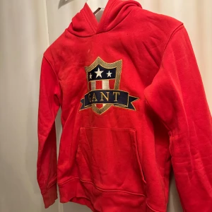 Röd hoodie från GANT - Säljer en snygg röd hoodie från GANT med ett stort broderat emblem på bröstet. Den har en klassisk känguruficka och är perfekt för både vardag och chill. Hoodien är i bra skick och superbekväm att ha på sig. Passar 10-12år.