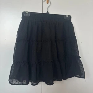 Svart volangkjol - Säljer en superfin svart kortkjol med volanger. Kjolen har ett elastiskt midjeband för en bekväm passform. Materialet är lätt och luftigt, knappt använd!