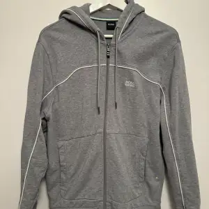 Säljer en stilren grå hoodie från Hugo Boss i mycket bra skick. Den har en dragkedja framtill och justerbar huva med snören. Den har vita detaljer längs sömmarna som ger en snygg kontrast.