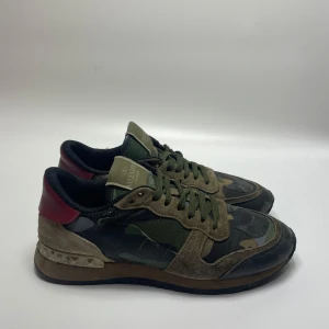 Valentino Rockrunner  - Säljer ett par snygga Valentino Rockrunner sneakers i camouflage. De har en cool mix av gröna och bruna nyanser med röda detaljer på hälen. Skorna har snörning och är i bra skick, perfekta för en avslappnad men stilren look. Liten defekt i hälen 