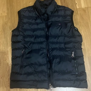 Svart dunväst från Tommy Hilfiger - Säljer en snygg svart dunväst från Tommy Hilfiger i storlek S. Västen har en dragkedja framtill och två praktiska fickor med dragkedjor. Perfekt för höst och vår när du vill hålla dig varm och stilren. 🖤