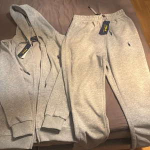 Grå hoodie och byxor från Polo Ralph Lauren. Storlek M men liten i storlek  - Säljer ett set med grå hoodie och matchande byxor från Polo Ralph Lauren. Hoodien har dragkedja och justerbar huva, medan byxorna har elastisk midja för extra komfort. Perfekt för en avslappnad stil eller träning. Båda plaggen är i nytt skick med etiketter kvar.