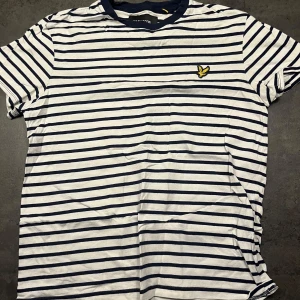 Randig t-shirt från Lyle & Scott - Säljer en snygg randig t-shirt från Lyle & Scott i vitt och marinblått. Den har en klassisk passform och korta ärmar, perfekt för sommaren. Detaljerad med en broderad gul logga på bröstet. Perfekt för en avslappnad stil! Nypris 200:-