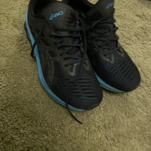 Svarta och blåa Asics löparskor - Säljer ett par Asics löparskor i svart med blå detaljer. Skorna har snörning och en bekväm sula som ger bra stöd vid löpning. Perfekta för träning och löprundor. Använda men i bra skick.