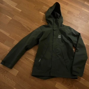 Grön regnjacka från Haglöfs - Säljer en grön regnjacka från Haglöfs i mycket bra skick. Jackan har en praktisk huva och dragkedja framtill. Perfekt för regniga dagar och utomhusaktiviteter. Den är långärmad och har en stilren design. Nypris ca 1500 kr