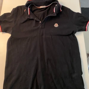 Svart pikétröja från Moncler - Säljer en snygg svart pikétröja från Moncler i bra skick. Den har korta ärmar och en klassisk krage med röd och vit randig detalj. Perfekt för både vardag och fest! 🖤