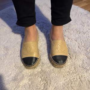 Säljer ett par snygga espadrillos från Chanel i beige och svart. De är gjorda av lammnappa och har en klassisk design med den ikoniska loggan på ovansidan. Perfekta för en stilren look under vår och sommar. Använda men helt okej sick 