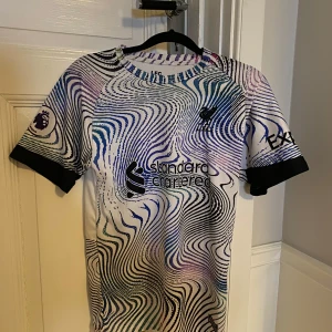 Liverpool FC fotbollströja - Säljer en cool Liverpool FC fotbollströja i vitt med ett psykedeliskt mönster i blått och lila. Tröjan är kortärmad och har tryck med 'M. Salah' och nummer 11 på ryggen. Perfekt för fans som vill sticka ut!