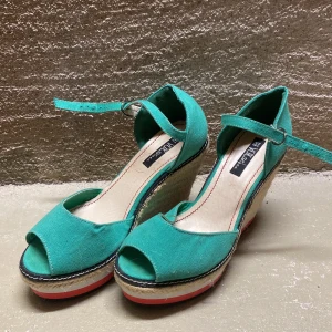 Turkosa kilklackar med ankelrem - Snygga turkosa kilklackar från Style Shoes med ankelrem och öppen tå. Skorna har en flätad kilklack och en bekväm passform, perfekta för sommarens fester. Sulan är i syntetmaterial och klacken ger en härlig höjd utan att kompromissa med komforten.