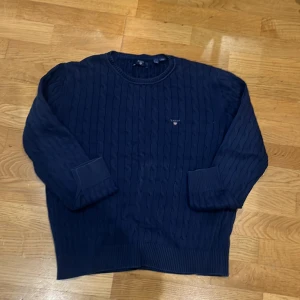 Mörkblå stickad tröja från GANT - Säljer en snygg mörkblå stickad tröja från GANT i bra skick. Den har ett klassiskt kabelstickat mönster och långa ärmar. Perfekt för höst och vinter! Tröjan är både stilren och bekväm, idealisk för både vardag och lite finare tillfällen. Fint skick, dock en liten vit fläck på baksida av armen, se bild. 
