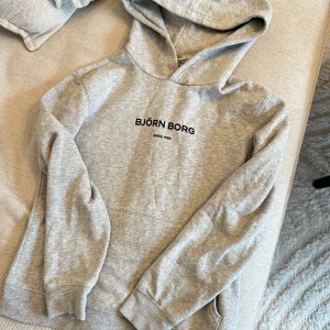 Grå hoodie från Björn Borg - Säljer en snygg och bekväm grå hoodie från Björn Borg. Perfekt för vardagsbruk med en klassisk design och svart text på bröstet. Den har en stor huva och är tillverkad i mjukt material för extra komfort. Passar perfekt till höst och vår!