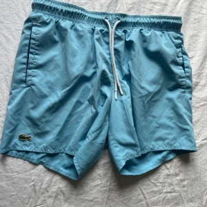 Ljusblå badshorts från Lacoste - Säljer ett par ljusblå badshorts från Lacoste i storlek M. De har en elastisk midja med vit snörning och sidofickor. Perfekta för sommardagar vid stranden eller poolen. 🏖️