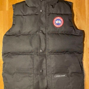 Svart dunväst från Canada Goose - Säljer en svart dunväst från Canada Goose i mycket bra skick. Västen har en broderad logga på bröstet och praktiska fickor framtill. Perfekt för kyliga höst- och vinterdagar. Passar både till vardags och utomhusaktiviteter.