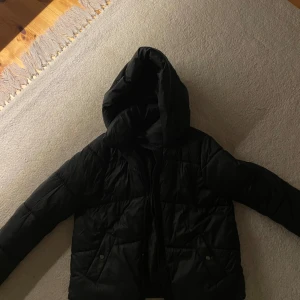Svart pufferjacka med huva - Säljer en svart pufferjacka i mycket bra skick. Den har en praktisk huva och långa ärmar, perfekt för kalla vinterdagar. Jackan är varm och stilren, idealisk för både vardag och utomhusaktiviteter. Passar perfekt för vintersäsongen!