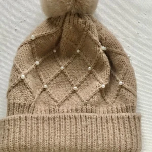Zara Beige stickad mössa med pärlor - Superfin beige stickad mössa med en fluffig boll på toppen. Den har ett snyggt rutmönster dekorerat med pärlor som ger en lyxig känsla. Perfekt för kalla vinterdagar och höstpromenader. Håller dig varm och stilren!