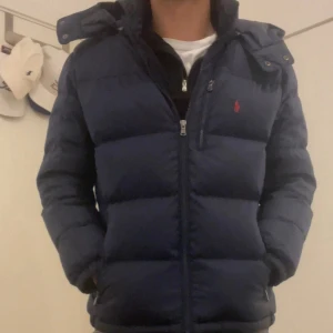 Mörkblå dunjacka från Ralph Lauren - Säljer en snygg mörkblå dunjacka från Ralph Lauren. Jackan har en dragkedja framtill och en liten röd logga på bröstet. Perfekt för kalla dagar med sin vadderade design och huva. Passar både till vardags och lite finare tillfällen. 🧥