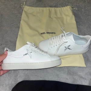 Vita sneakers från Axel Arigato - Säljer ett par stilrena vita sneakers från Axel Arigato i skinn. De har en snygg fågelbrodering på sidan och klassisk snörning. Perfekta för både vardag och fest! Skorna är i mycket bra skick och kommer med originalpåse. Säljer då de är fel storlek på dom är en 38 i vanligt fall men dessa är i 36. Nypris på skorna är 2500. Har använt fom i 30 minuter bara sen dess har dom Sldig blivit använda igen.