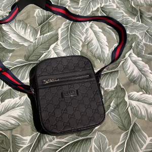 Svart axelväska från Gucci - Säljer en snygg svart axelväska från Gucci med det klassiska GG-mönstret. Väskan har en justerbar axelrem i rött och blått, samt en praktisk dragkedja framtill. Perfekt för både vardag och fest! Hör av dig om du har några frågor. Pris är sänkbart.