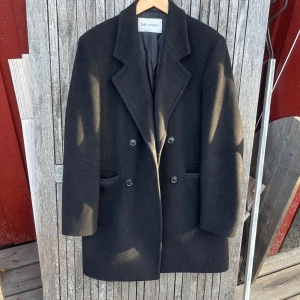 Svart kappa från Our Legacy - Säljer en stilren svart kappa från Our Legacy i storlek 46. Gjord av mjuk ullblandning med 70% virgin ull, 20% polyamid och 10% kashmir. Perfekt för höst och vinter med sin klassiska design. Kappan har en dubbelknäppt front och är i mycket bra skick. 
