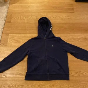 Mörkblå hoodie från Ralph Lauren - Säljer en snygg mörkblå hoodie från Ralph Lauren i storlek 14-16år. Den har en dragkedja framtill och en broderad logga på bröstet. Perfekt för höst och vår, med långa ärmar och en bekväm passform. Huvan har texten 'POLO' tryckt på sig. Passar både till vardags och chillkvällar.