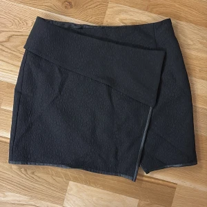 Omlottkjol - Jättesnygg svart omlottkjol i storlek S. Kjolen har en stilren design med en omlott-detalj som ger en modern touch. Kjolen har lågmidja och är kort.