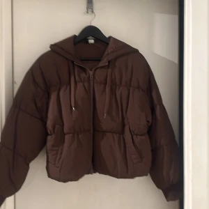 Brun pufferjacka med huva - Säljer en brun pufferjacka i mycket bra skick. Den är perfekt för höst och vinter med sin varma och mysiga design. Jackan har en dragkedja framtill och en praktisk huva med snörning. Den är croppad och har långa ärmar, vilket ger en modern look. Perfekt för kyliga dagar!