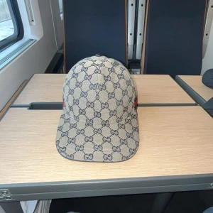 Gucci keps  - Säljer en snygg Gucci keps. Kepsen är i beige färg med blåa detaljer. Kepsen är i storlek (S) Du kan justera kepsen till din storlek. Kepsen är (ÄKTA) Kepsen är köpt för ca 1 år sedan. . Kan tänka mig byta mot andra saker!