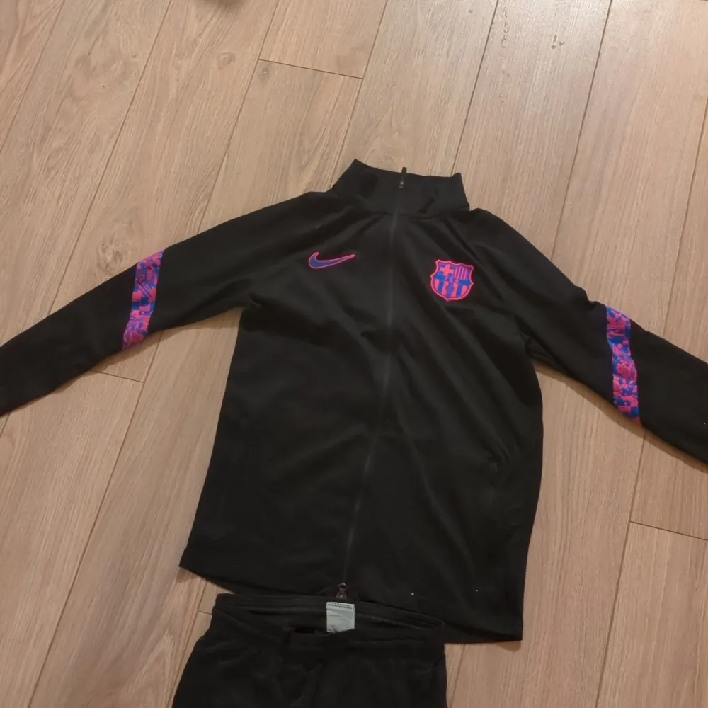 Säljer ett svart träningsset från Nike med Barcelona-logga. Setet har en jacka med dragkedja och matchande byxor. Detaljer i lila och rosa på ärmar och ben ger en cool look. Perfekt för fotbollsträning eller vardagsbruk. Dri-FIT material för bästa komfort.Finns ett lite hål i byxan med det syns knappt. Storlek L barn 147-157. Tröjor & Koftor.