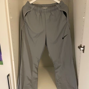 Gråa trackbyxor från Nike - Säljer ett par grå Nike Dri-FIT träningsbyxor i storlek M. De har en elastisk midja och är perfekta för träning eller att klä ut dom. Byxorna är helt nya och har en snygg svart Nike-logga på sidan. Perfekta fö både löprundan och stan