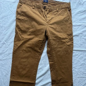 Bruna chinos från GANT - Säljer ett par snygga bruna chinos från GANT i bra skick. De har en normal passform och är perfekta för både vardag och lite finare tillfällen. Byxorna har knappar och bälteshällor, vilket gör dem både praktiska och stiliga. Perfekta för höst och vår!