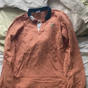 Orange pikétröja med broderad detalj. U.S POLO ASSN - Säljer en snygg orange pikétröja med en broderad detalj på bröstet. Tröjan har en klassisk krage i mörkblått och vita knappar. Perfekt för en avslappnad stil. Passar bra till både jeans och chinos!