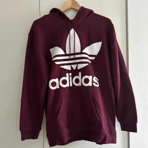 Vinröd hoodie från Adidas - Säljer en snygg vinröd hoodie från Adidas med det klassiska logotyptrycket i vitt på framsidan. Den har en stor ficka fram och en bekväm huva. Perfekt för höst och vinter, både mysig och stilren. Passar till alla tillfällen!
